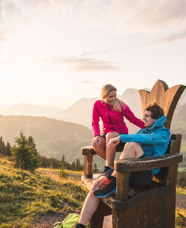 Couple enjoys sunset while hiking, seated on giant wooden throne with scenic view over Hochkönig valley and mountain peaks | © Hochkönig Tourismus GmbH / Christian Schartner
